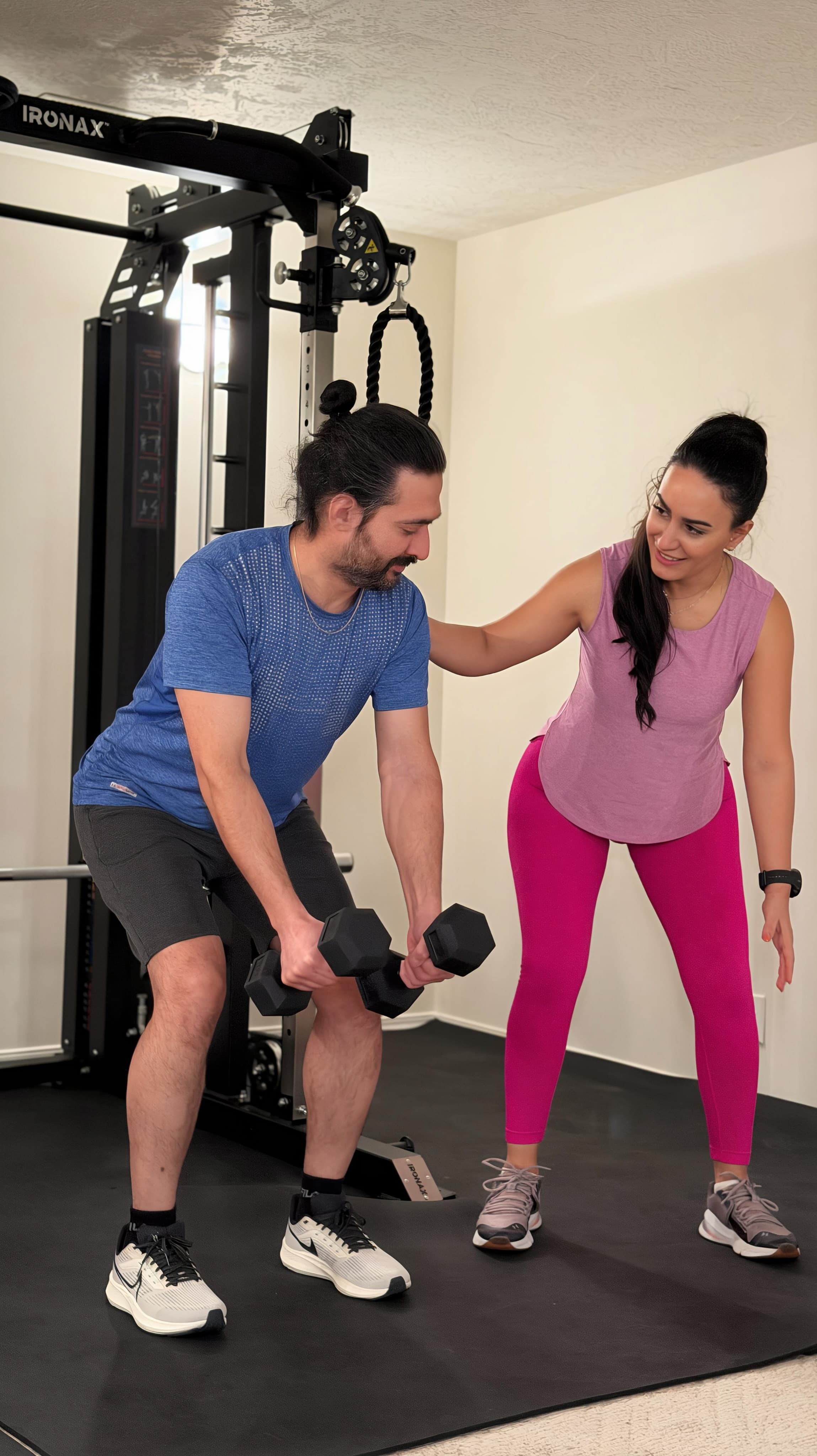 Mahnaz coaching a client through a dumbbell movement beside cable machine equipment.